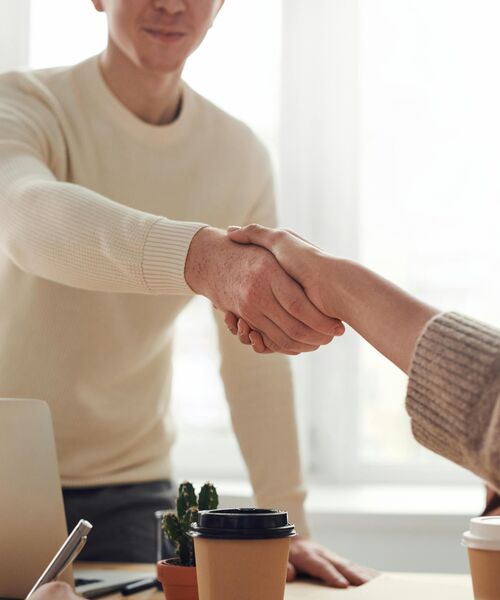 Zwei Personen geben sich die Hand in einer hellen Büroumgebung, auf dem Tisch stehen Kaffeebecher und ein Laptop.
