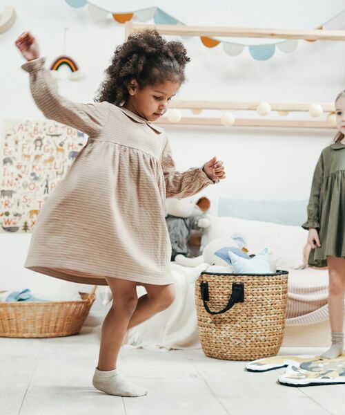 Ein Kind tanzt fröhlich in einem hell eingerichteten Kinderzimmer, während ein weiteres Kind im Hintergrund steht und zusieht.