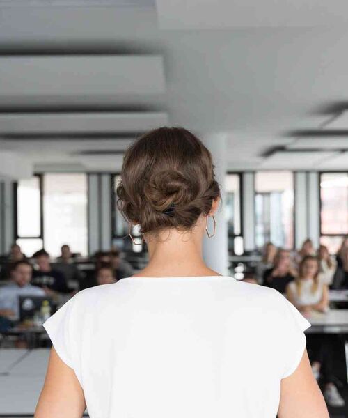 Frau steht vorne im Raum und spricht vor einer Gruppe von Zuhörerinnen und Zuhörern.