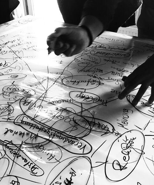 Schwarz-weiß-Foto von Händen, die über ein großes Blatt Papier mit vielen handgeschriebenen Notizen und Kreisen arbeiten – eine kreative oder planerische Gruppensituation.