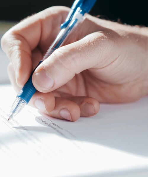 Eine Hand hält einen blauen Kugelschreiber und unterschreibt ein Dokument auf weißem Papier.