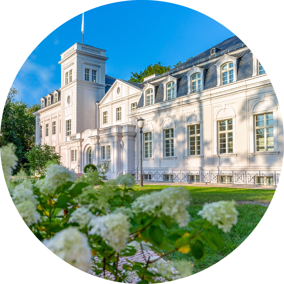 Historisches weißes Gebäude mit Turm und Garten, im Vordergrund blühende Sträucher bei sonnigem Wetter.