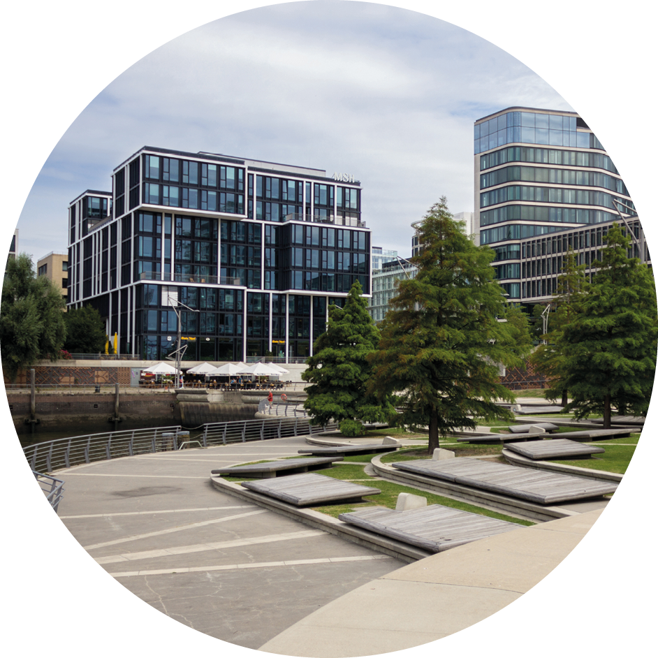 Modernes Bürogebäude mit Glasfassade an einem Kanal, umgeben von Bäumen und Sitzflächen im Parkbereich.